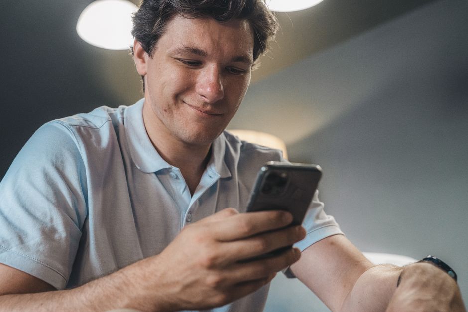 A man in a blue polo shirt smiling while using a smartphone indoors.